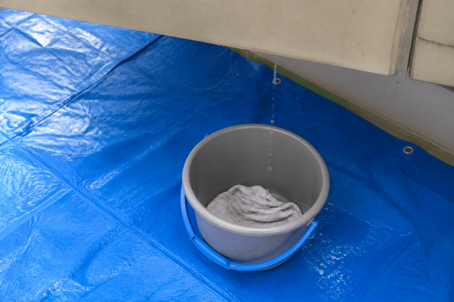 ブルーシートを敷いた床に置かれたバケツ。水滴が落ちる位置に置かれ、中には水はね防止用の雑巾が敷かれている様子。DIY初心者でも真似しやすい、具体的な設置例の写真。]