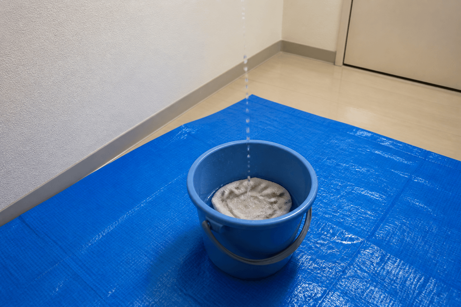 実際に天井からの水滴をバケツで受けている様子。バケツの中に雑巾が入っており、床には水はね防止のブルーシートが敷かれている状況がわかる写真。]